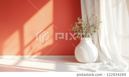 White vase with green leaves sits on a table in front of a red wall. The vase is the only object in the room, and it is the focal point of the space. The red wall and white curtains create a warm 122834269