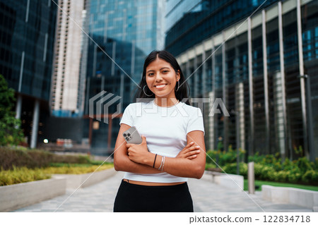 Confident Young Woman in Modern Urban Setting Smiling Happily Amidst Surrounding Buildings Confident Young Woman in Modern Urban Setting Smiling Happily Amidst Surrounding Buildings 122834718