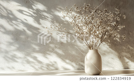 White vase with flowers in it sits on a table. The vase is filled with white flowers, and the sunlight is shining on it, creating a warm and inviting atmosphere 122834919