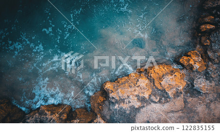 Rocky shoreline with a body of water in the background. The water appears to be a mix of blue and green, with some white foam on the surface. The rocks are scattered throughout the scene Rocky shoreline with a body of water in the background. The water appears to be a mix of blue and green, with some white foam on the surface. The rocks are scattered throughout the scene 122835155