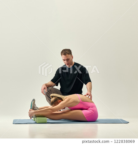 Trainer assists woman in pink sportswear performing deep forward stretch on yoga mat, ensuring correct posture and flexibility against studio background. Trainer assists woman in pink sportswear performing deep forward stretch on yoga mat, ensuring correct posture and flexibility against studio background. 122838069