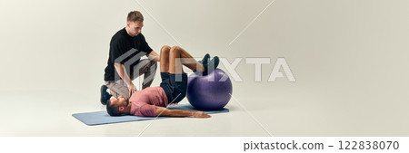 Trainer guiding man in bridge exercise with stability ball, ensuring controlled movement for recovery against light studio background. Trainer guiding man in bridge exercise with stability ball, ensuring controlled movement for recovery against light studio background. 122838070