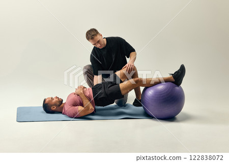 Trainer guiding man in bridge exercise with stability ball, ensuring controlled movement for recovery against light studio background. Trainer guiding man in bridge exercise with stability ball, ensuring controlled movement for recovery against light studio background. 122838072
