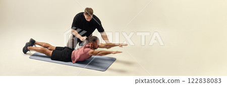 Trainer correcting man form in back extension exercise, stabilizing movement for recovery against white studio background. 122838083