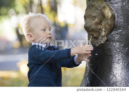 The kid washes his hands in the fountain. The boy put his fingers under the stream of water in the park 122840164