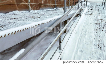 Close-up of a ramp with metal railings, covered with snow, for baby strollers and handicapped accessibility Close-up of a ramp with metal railings, covered with snow, for baby strollers and handicapped accessibility 122842657