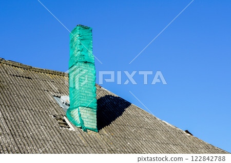 A house chimney covered with a green safety net on a gray damaged slate roof against a blue sky A house chimney covered with a green safety net on a gray damaged slate roof against a blue sky 122842788