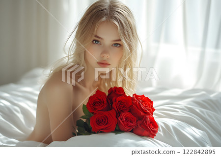 happy blonde woman with blue eyes holding red roses, lying on a white bed in soft natural light, celebrating a special occasion 122842895