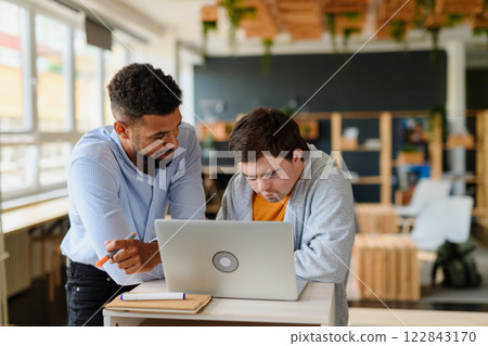 Happy young man with Down syndrome and his tutor using lapotp indoors at school. 122843170