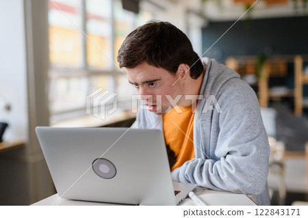 Concentrated young man with Down syndrome sitting and studying indoors at school, using laptop Concentrated young man with Down syndrome sitting and studying indoors at school, using laptop 122843171