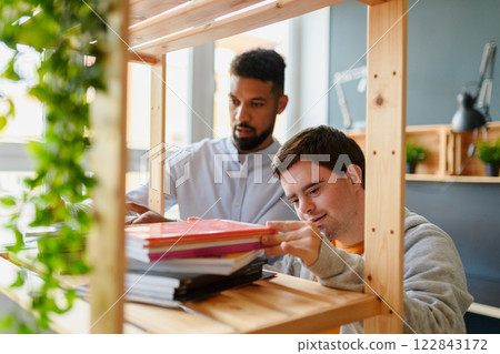 Young happy man with Down syndrome and his tutor choosing books for reading. Young happy man with Down syndrome and his tutor choosing books for reading. 122843172