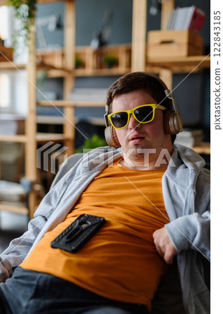 Close up of happy young man with Down syndrome with sunglasses and headphones sitting indoors 122843185