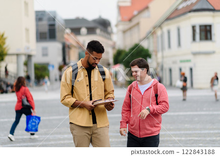 A young man with Down syndrome and his mentoring friend walking across city, talking. A young man with Down syndrome and his mentoring friend walking across city, talking. 122843188