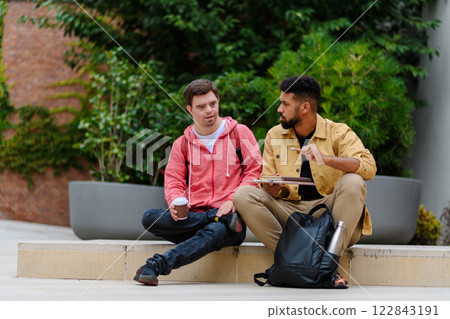 Happy young man with Down syndrome and mentoring friend sitting and talking outdoors, spending time together. 122843191