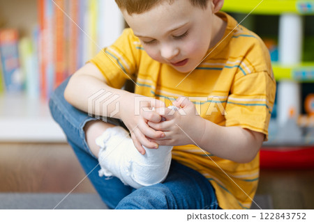 A little toddler boy holds his leg in a cast with his hands. Fracture of a foot and finger in children. Human healthcare and medicine concept. Bandage 122843752