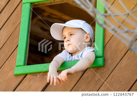 Pensive toddler boy of three or four years looks out of a window on a wooden playground. Toddlerhood and childhood concept. Active lifestyle Pensive toddler boy of three or four years looks out of a window on a wooden playground. Toddlerhood and childhood concept. Active lifestyle 122843767
