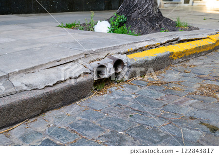 A cracked sidewalk, yellow curb, and a tree in the back 122843817