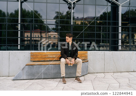 Wide shot of handsome young man in casual clothing sitting on modern bench in city street, holding two disposable coffee cups, enjoying break in front of modern building. Concept of urban lifestyle. 122844941