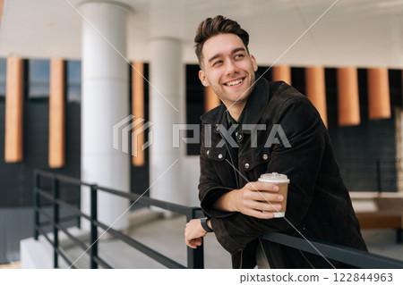 Handsome positive young man relaxing during coffee break, leaning on railing standing by modern office building. Happy bearded male in casual attire smiling looking away. Concept of urban lifestyle. 122844963