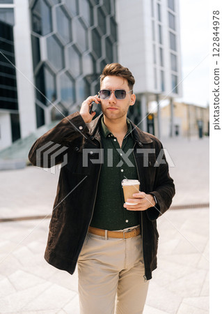 Vertical portrait of stylish young man in sunglasses with serious expression having phone call while holding takeaway coffee standing outside office building. Concept of successful city lifestyle. Vertical portrait of stylish young man in sunglasses with serious expression having phone call while holding takeaway coffee standing outside office building. Concept of successful city lifestyle. 122844978