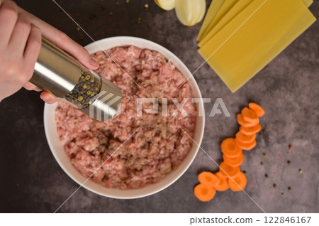 Top view raw ground minced meat in white bowl, woman's hands holding pepper mill. Lasagne pasta sheets, onion. carrot, pepper. Black background. Homemade lasagna cooking preparation Top view raw ground minced meat in white bowl, woman's hands holding pepper mill. Lasagne pasta sheets, onion. carrot, pepper. Black background. Homemade lasagna cooking preparation 122846167