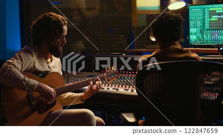 Skilled songwriter guitarist playing musical instrument in professional studio, recording tracks and editing them on mixing console. Artist musician singing and producing music. Camera A. 122847659