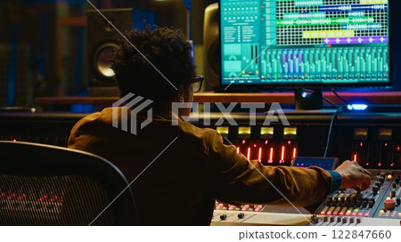 Sound designer uses mixing console and managing audio tracks, twisting knobs on control desk for adjusting tunes and volume. Producer creates music in professional studio, technical gear. Camera A. 122847660