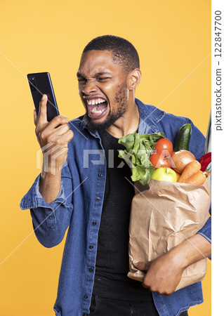 African american irritated guy screaming at his smartphone during fight with a friend, getting angry and mad on remote phone call. Adult with groceries shouting at someone. African american irritated guy screaming at his smartphone during fight with a friend, getting angry and mad on remote phone call. Adult with groceries shouting at someone. 122847700