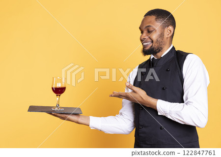 Restaurant worker serving people with a glass of red wine before dinner, recommending a special drink for customer satisfaction. Waiter carrying a tray for a taste test, professionalism. 122847761