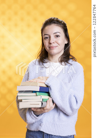 Portrait of smiling woman holding pile of books, conveying joy of reading concept, isolated over studio background. Cheerful person with stack of novels showing appreciation for literature 122847764
