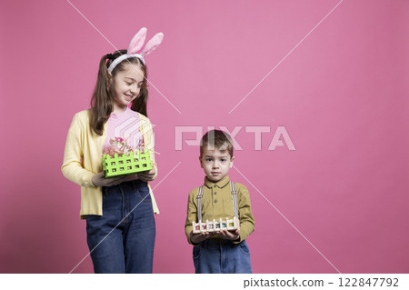 Smiling cute siblings showing festive colorful decorations on camera, being excited about easter celebration and spring time. Brother and sister presenting their baskets filled with painted eggs. Smiling cute siblings showing festive colorful decorations on camera, being excited about easter celebration and spring time. Brother and sister presenting their baskets filled with painted eggs. 122847792