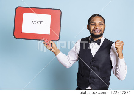Professional front desk staff presenting a red speech bubble with a vote message, pushing guests into politics. Five star hotel personnel showing the billboard icon for an ad commercial. Professional front desk staff presenting a red speech bubble with a vote message, pushing guests into politics. Five star hotel personnel showing the billboard icon for an ad commercial. 122847825