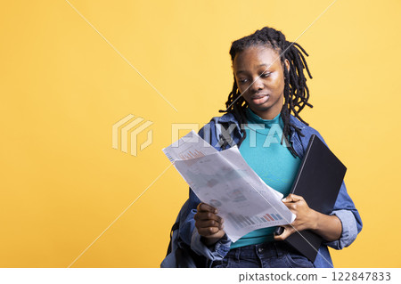African american woman working accounting job, looking at financial report pages, studio background. Young girl reading paperwork containing economic graphs and figures, solving bookkeeping tasks 122847833