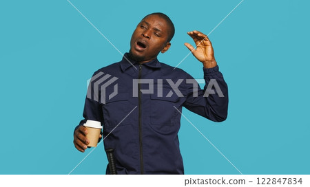 Portrait of fatigued security guard drinking coffee cup to remain energized while overseeing premises. Private law enforcement worker consuming caffeinated beverage, studio backdrop, camera A 122847834