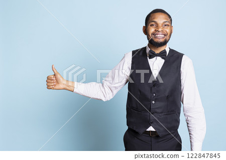 Smiling bellboy in suit and tie showing thumbs up gesture in studio, offering his approval and being satisfied. Positive doorkeeper gives a like sign, trust and professionalism. 122847845