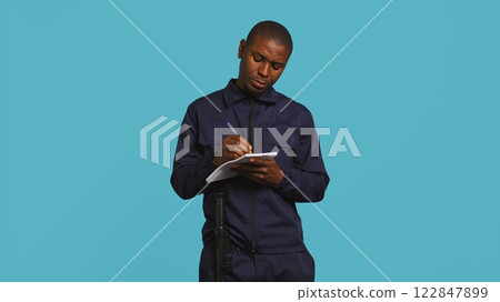 Security guard with notepad taking notes, receiving orders about suspects to look out. Portrait of bodyguard using pen, documenting layout location in need of protection, studio background, camera B 122847899