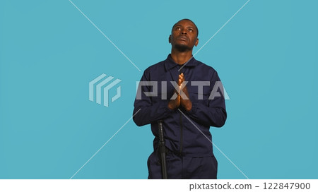Portrait of security guard waiting for miracle, putting hands together while overseeing premises. Private law enforcement worker looking to sky, praying, studio backdrop, camera B 122847900
