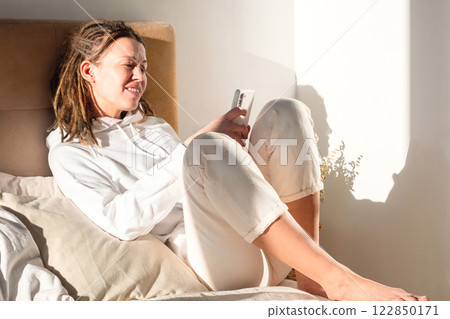 Woman Using Smartphone on Sunlit Bed. Relaxation and Modern Lifestyle. 122850171