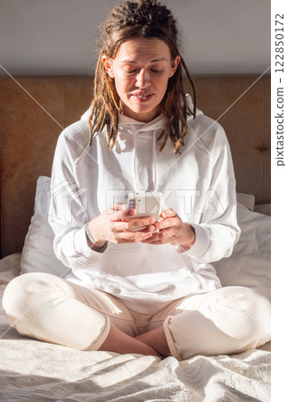 Woman Using Smartphone on Sunlit Bed. Relaxation and Modern Lifestyle. 122850172