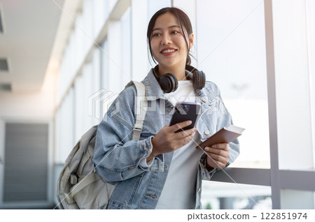 A positive Asian female traveler stands by a window in the airport, smiling as she daydreams. A positive Asian female traveler stands by a window in the airport, smiling as she daydreams. 122851974