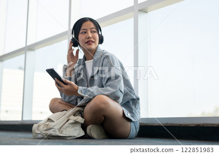 A beautiful Asian female passenger sits on the floor in the airport terminal, enjoying music. A beautiful Asian female passenger sits on the floor in the airport terminal, enjoying music. 122851983