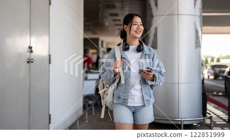 A positive, smiling Asian female traveler walks outside the airport, arriving at her destination. 122851999