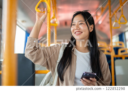 A cheerful Asian woman holds a handrail while admiring the city view during her bus ride. 122852298