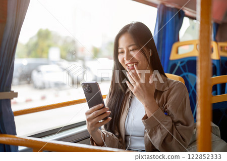 A cheerful Asian woman enjoys chatting on her smartphone while on a bus, commuting in the city. 122852333