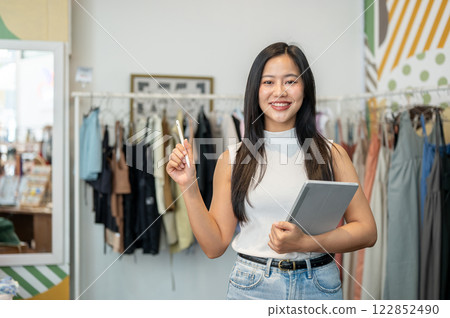 A confident Asian female designer stands in her shop with a digital tablet in her hand, smiling. A confident Asian female designer stands in her shop with a digital tablet in her hand, smiling. 122852490