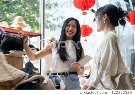 A smiling Asian shop employee is recommending new items to a customer, helping them select items. A smiling Asian shop employee is recommending new items to a customer, helping them select items. 122852529
