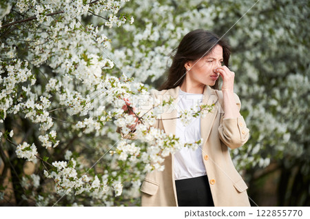 Woman allergic suffering from seasonal allergy at spring in blossoming garden at springtime. Young woman sneezing, having runny nose in front of blooming tree. Spring allergy concept Woman allergic suffering from seasonal allergy at spring in blossoming garden at springtime. Young woman sneezing, having runny nose in front of blooming tree. Spring allergy concept 122855770