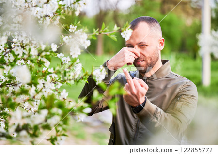 Man allergic suffering from seasonal allergy at spring in blossoming garden at springtime. Man showing middle finger gesture, having runny nose in front of blooming tree. Spring allergy concept. Man allergic suffering from seasonal allergy at spring in blossoming garden at springtime. Man showing middle finger gesture, having runny nose in front of blooming tree. Spring allergy concept. 122855772