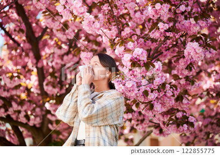 Woman allergic suffering from seasonal allergy at spring in blossoming garden at springtime. Woman sneezing and blowing nose using nasal handkerchief in front of blooming tree. Spring allergy concept. 122855775