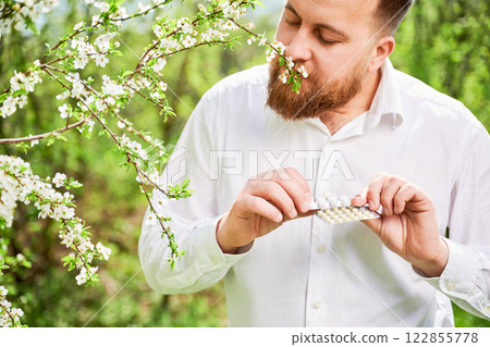 Man allergic suffering from seasonal allergy at spring. Handsome man holding pack of pills, sniffing flowers in blossoming garden at springtime. Antihistamine medication concept 122855778
